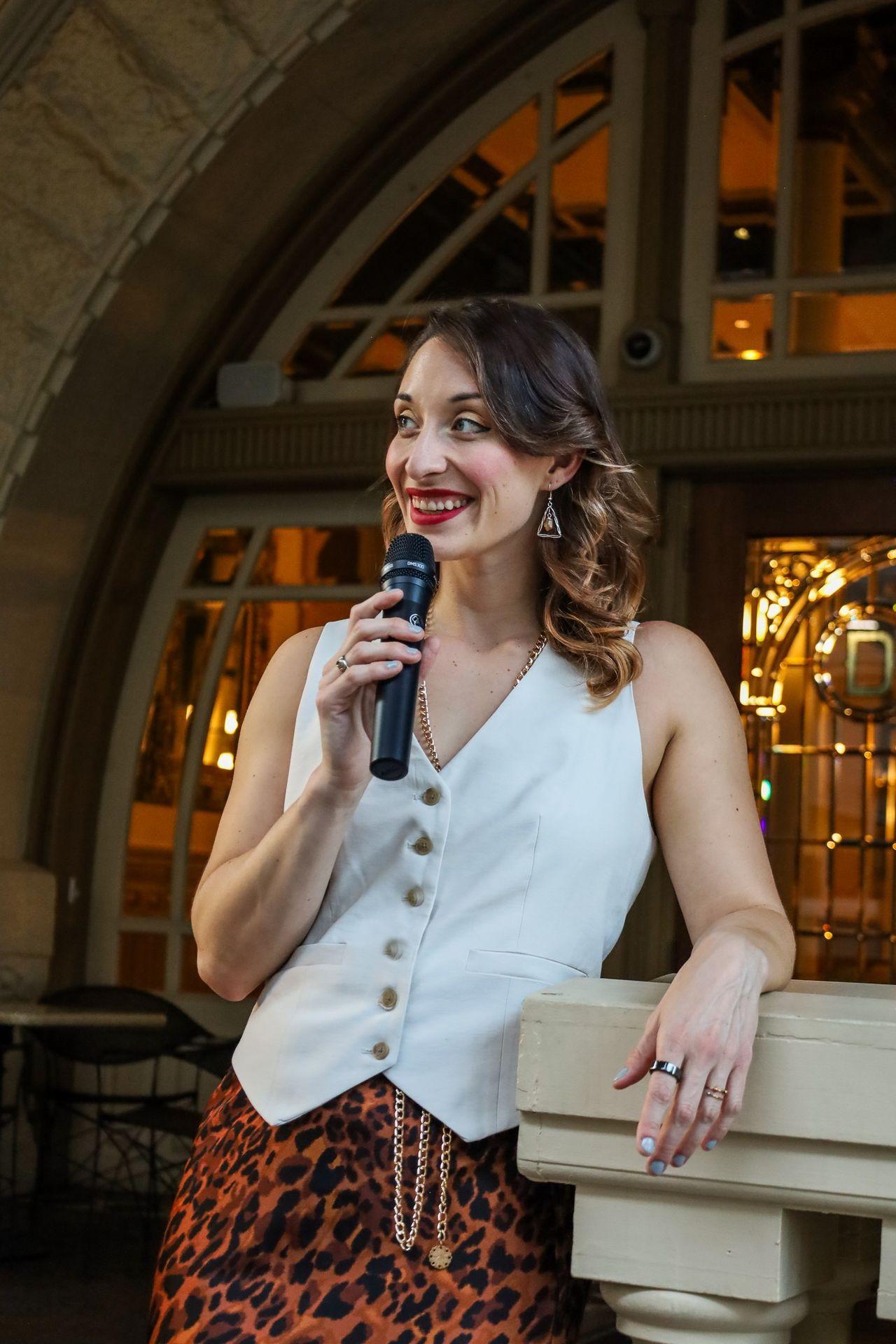 Woman in a white vest and leopard skirt holding a microphone, standing by an ornate entrance.