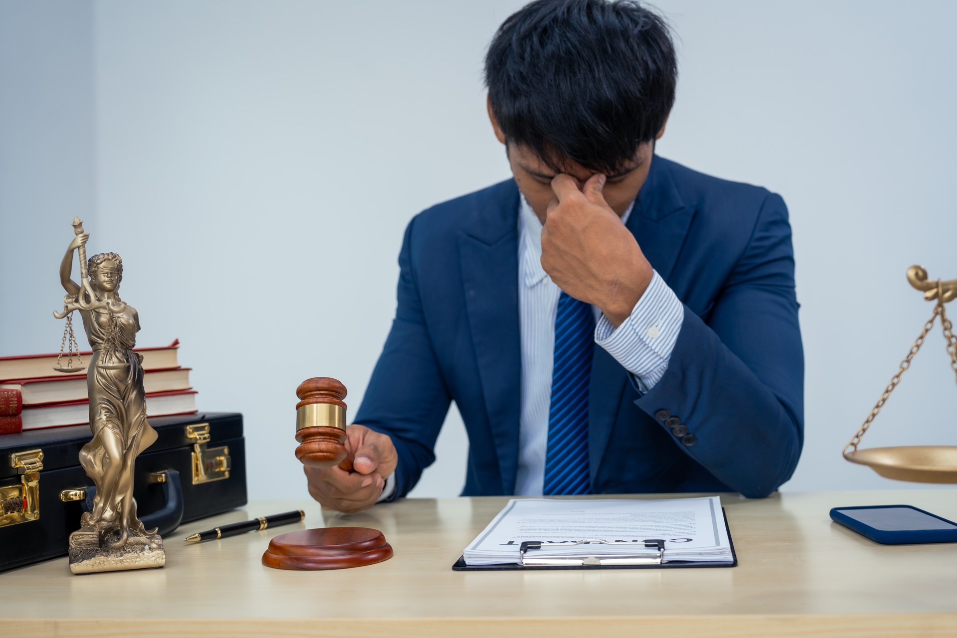 An Asian businessman in a formal suit works alone at a lawyer's desk, by golden scales, law books, leather bags, and a Lady Justice statue, emphasizing personal freedom and legal empowerment.